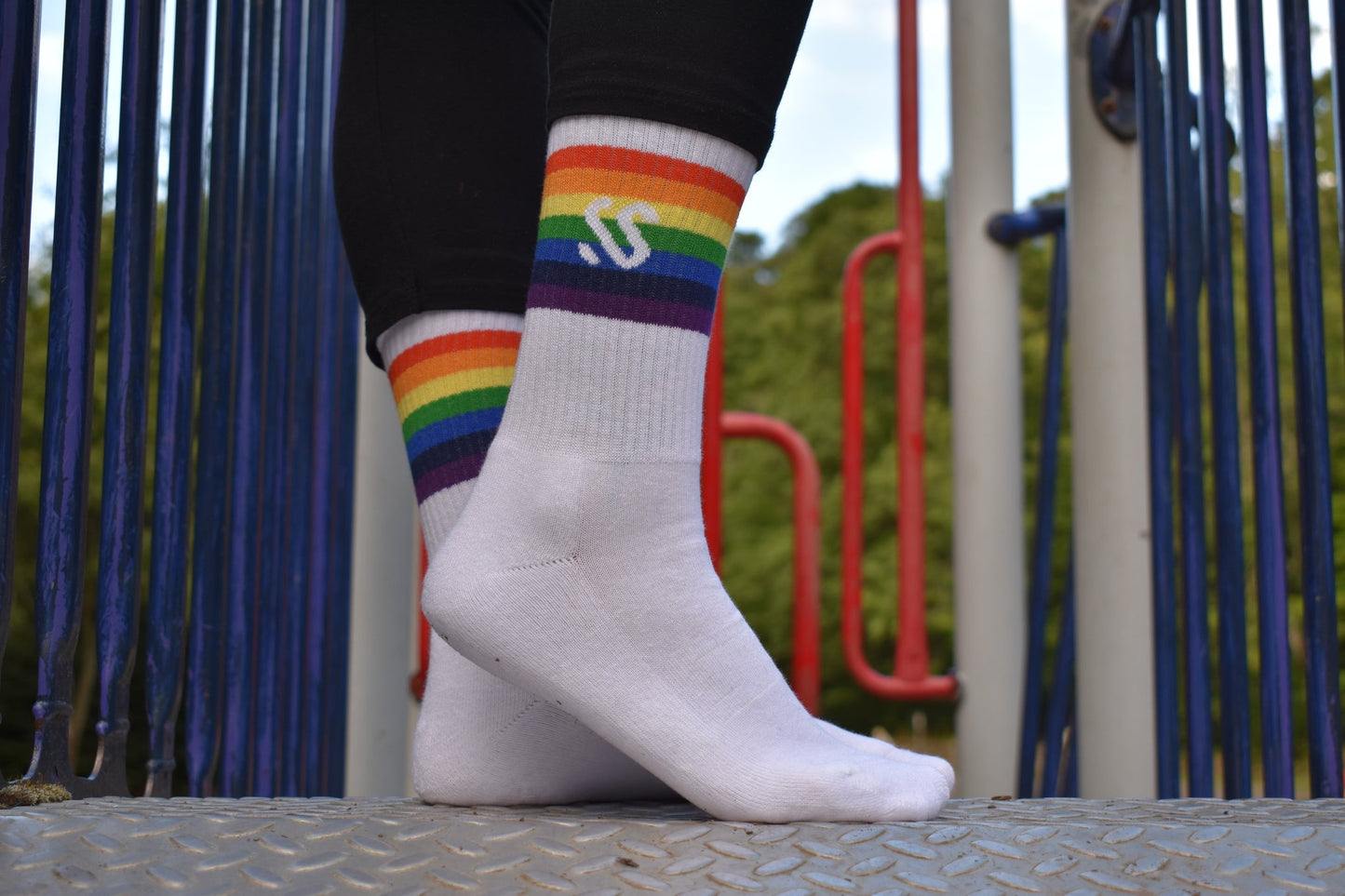 A pair of white crew length socks with a rainbow band being worn outside